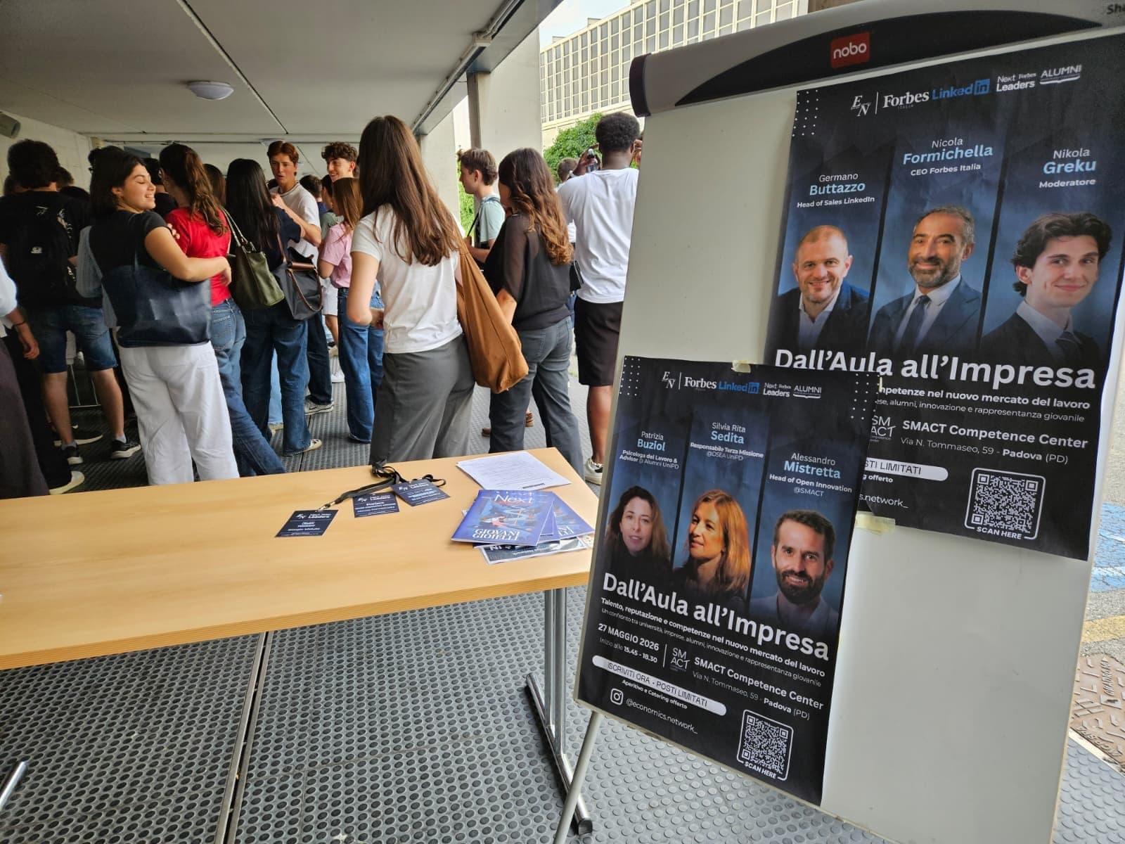 Due locandine dell'incontro «Dall'Aula all'Impresa» su un cavalletto, accanto al tavolo della registrazione e agli studenti in attesa