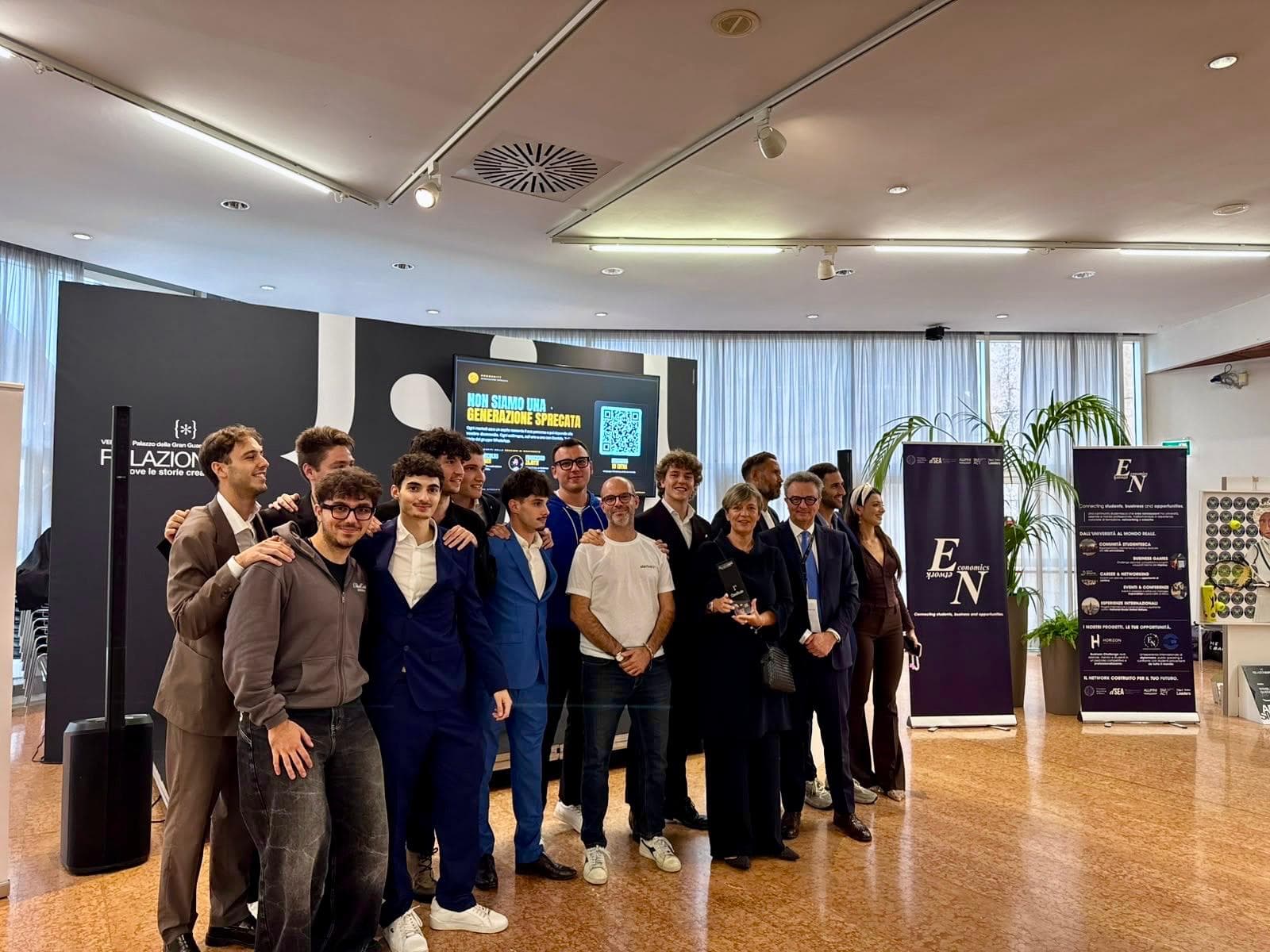 Foto di gruppo della premiazione: il team di Economics Network con giuria e rappresentanti delle istituzioni, davanti allo schermo di RelazionEXPO