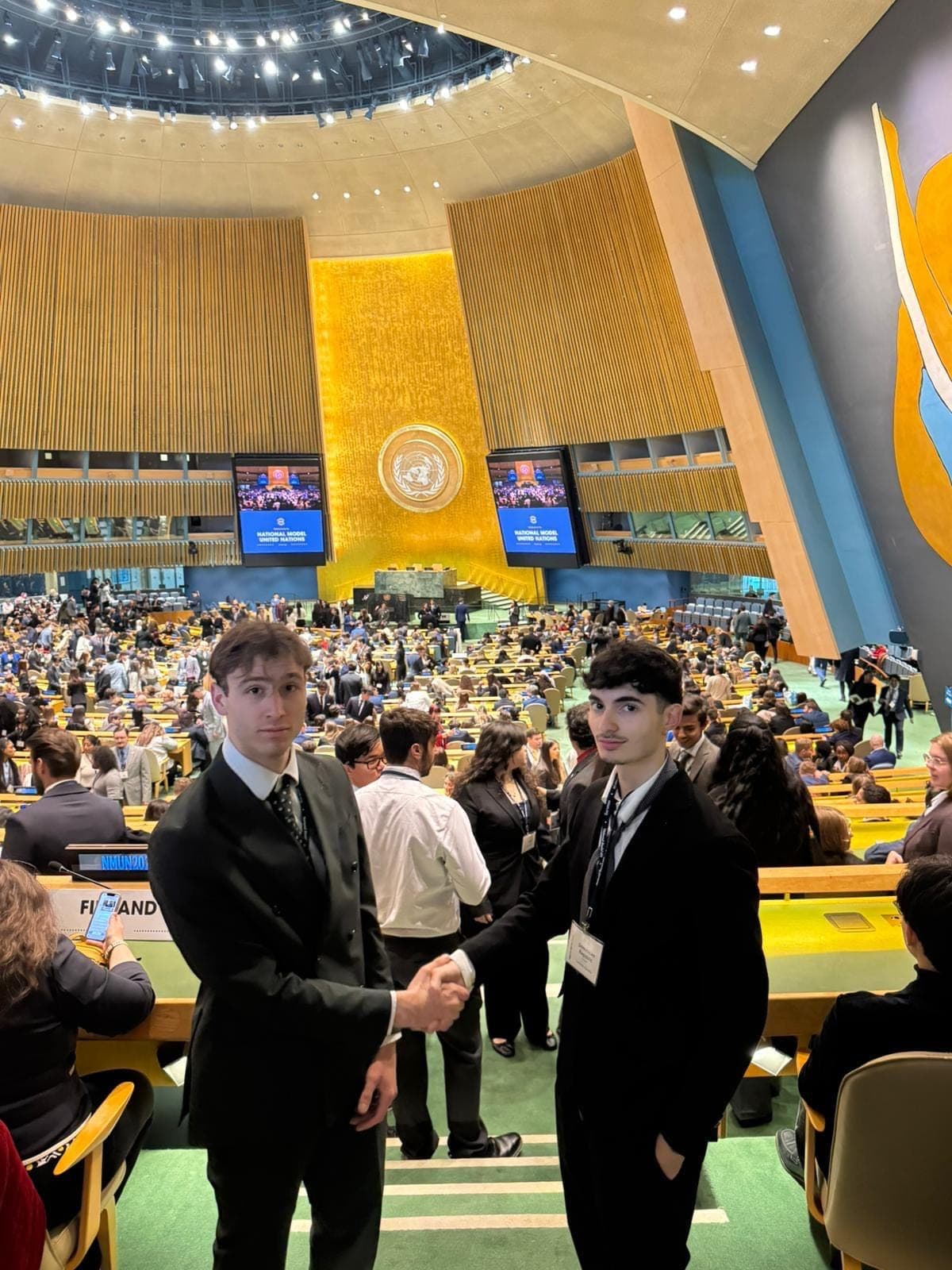 Due ragazzi in abito scuro si stringono la mano in una grande sala assembleare gremita di studenti; sugli schermi la scritta National Model United Nations